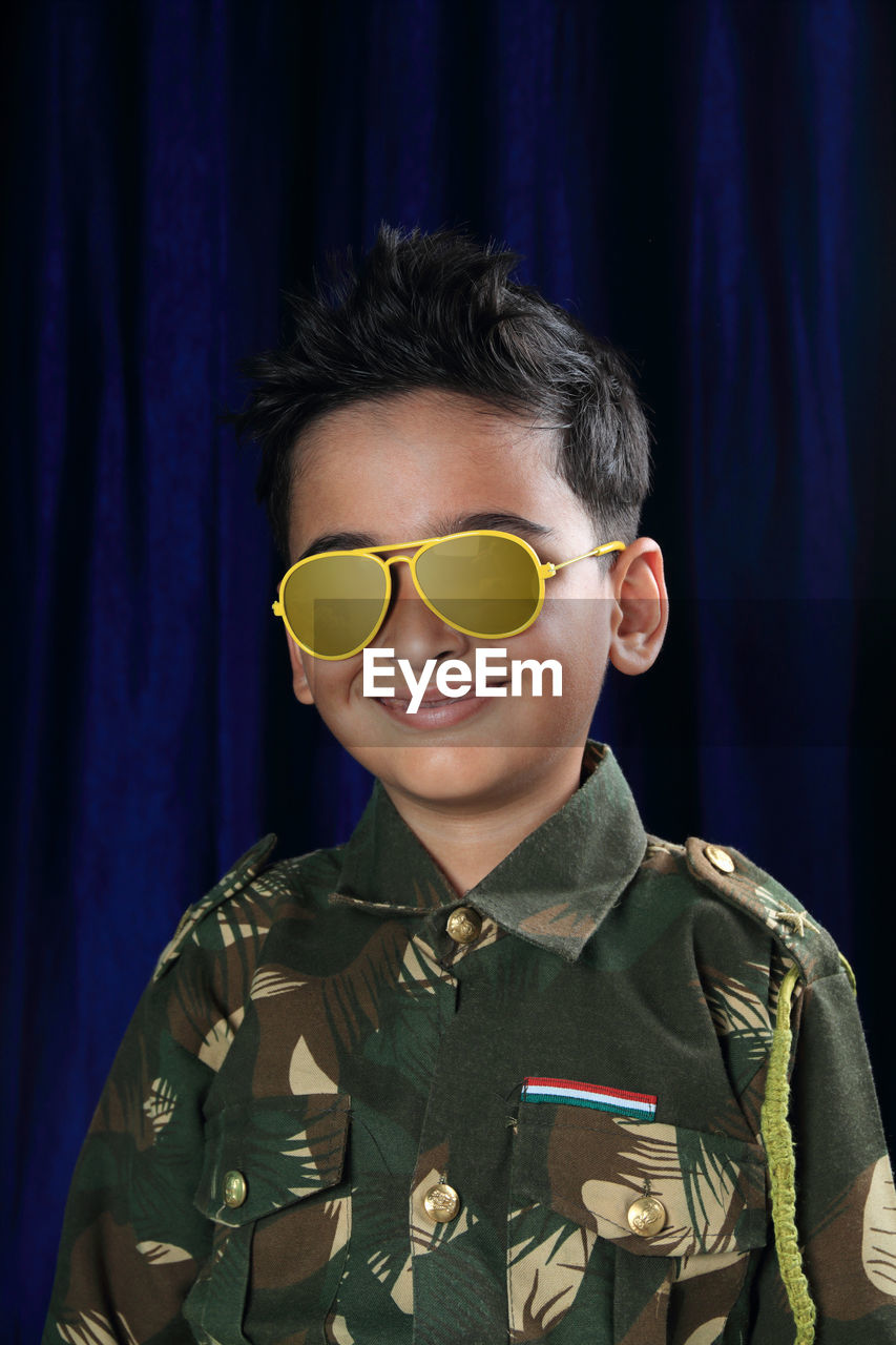 Portrait of smiling boy in army uniform and sunglasses standing against curtain