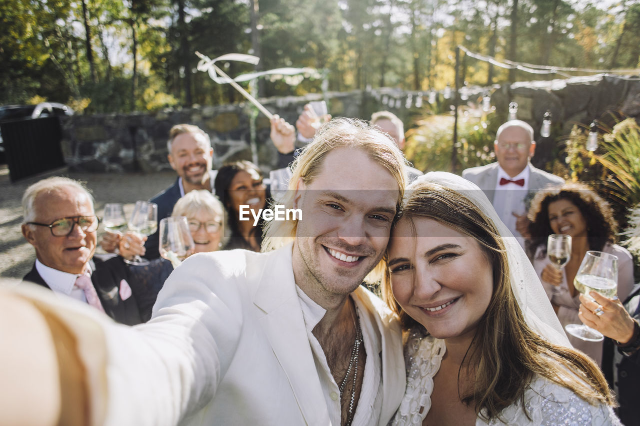 Portrait of happy newlywed couple taking selfie with guests in background on wedding day