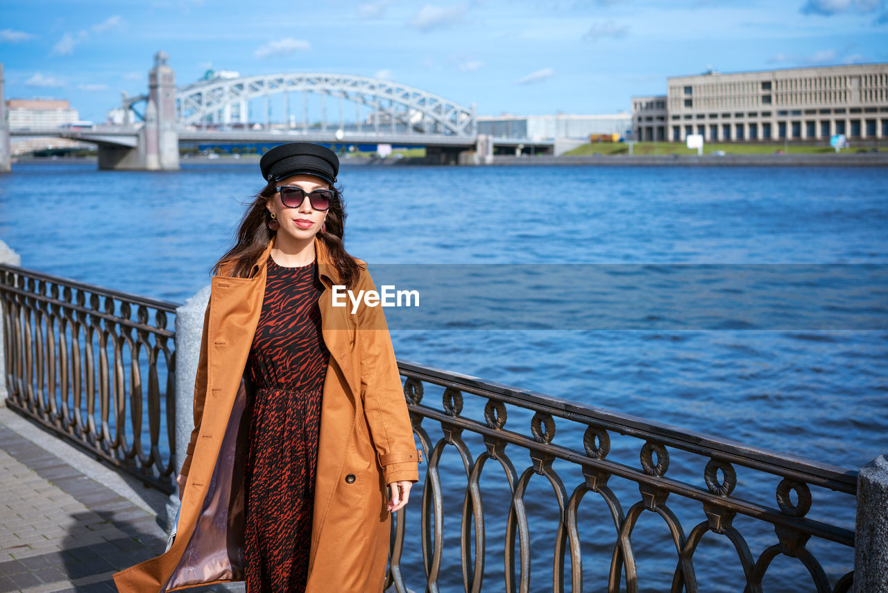 Young woman in sunglasses posing on embankment in brown coat on spring sunny day