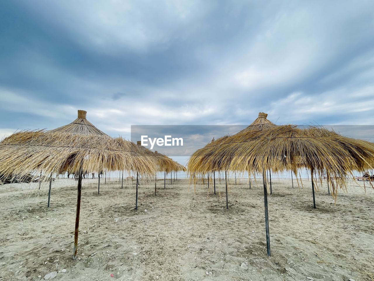 thatched roof, land, sky, cloud, roof, sand, nature, parasol, umbrella, beach, sea, beauty in nature, beach umbrella, tranquility, scenics - nature, tranquil scene, no people, shade, hut, sunshade, protection, day, environment, travel destinations, outdoors, landscape, thatching, idyllic, water, vacation, holiday, security, trip, summer, tropical climate, travel, straw, non-urban scene, shore, absence, ocean, chair, sunlight