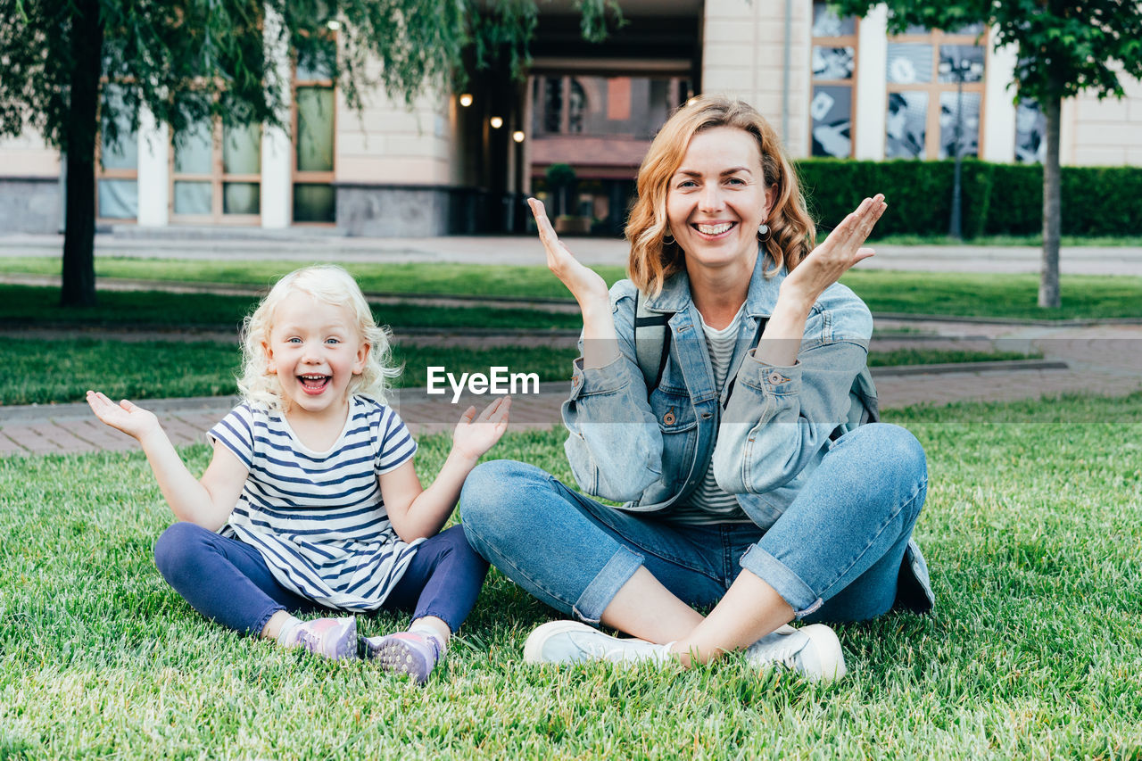 Mom and her little daughter are sitting on the lawn playing and having fun looking at the camera.