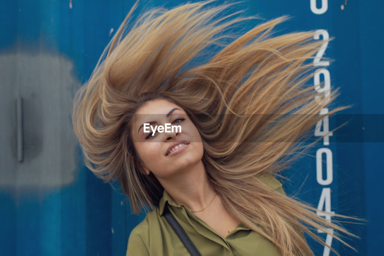 Portrait of smiling young woman tossing hair against blue metal