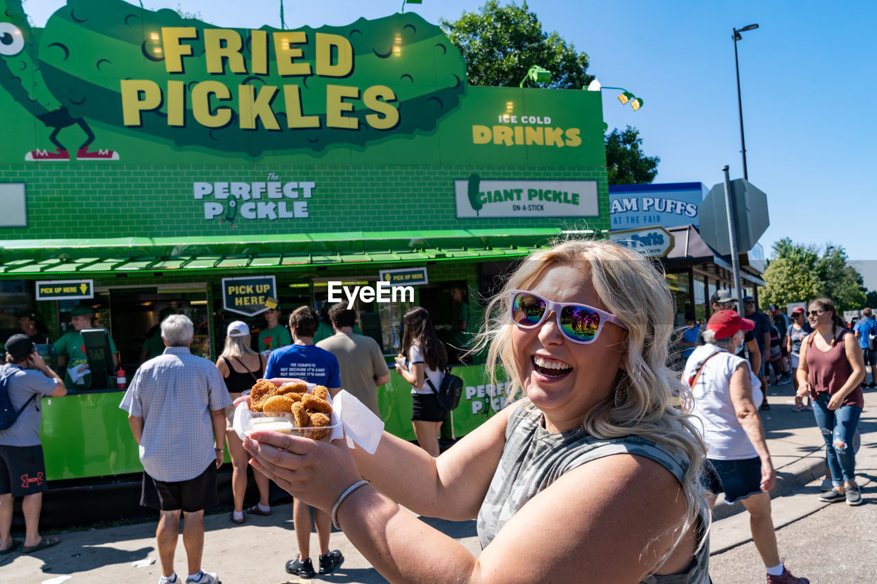 PORTRAIT OF SMILING WOMAN WITH SUNGLASSES AGAINST PEOPLE IN BACKGROUND