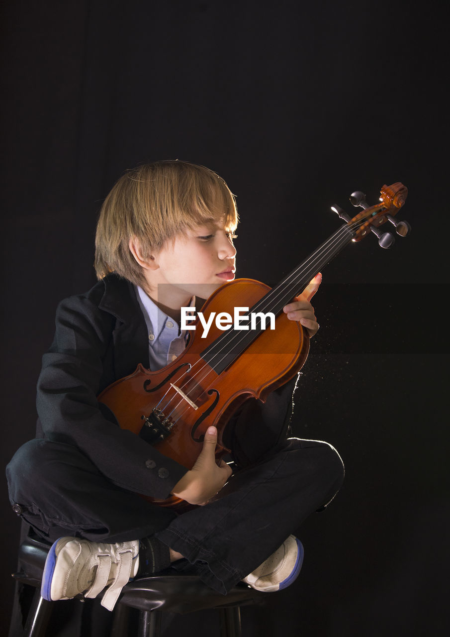 Boy holding violin while sitting against black background