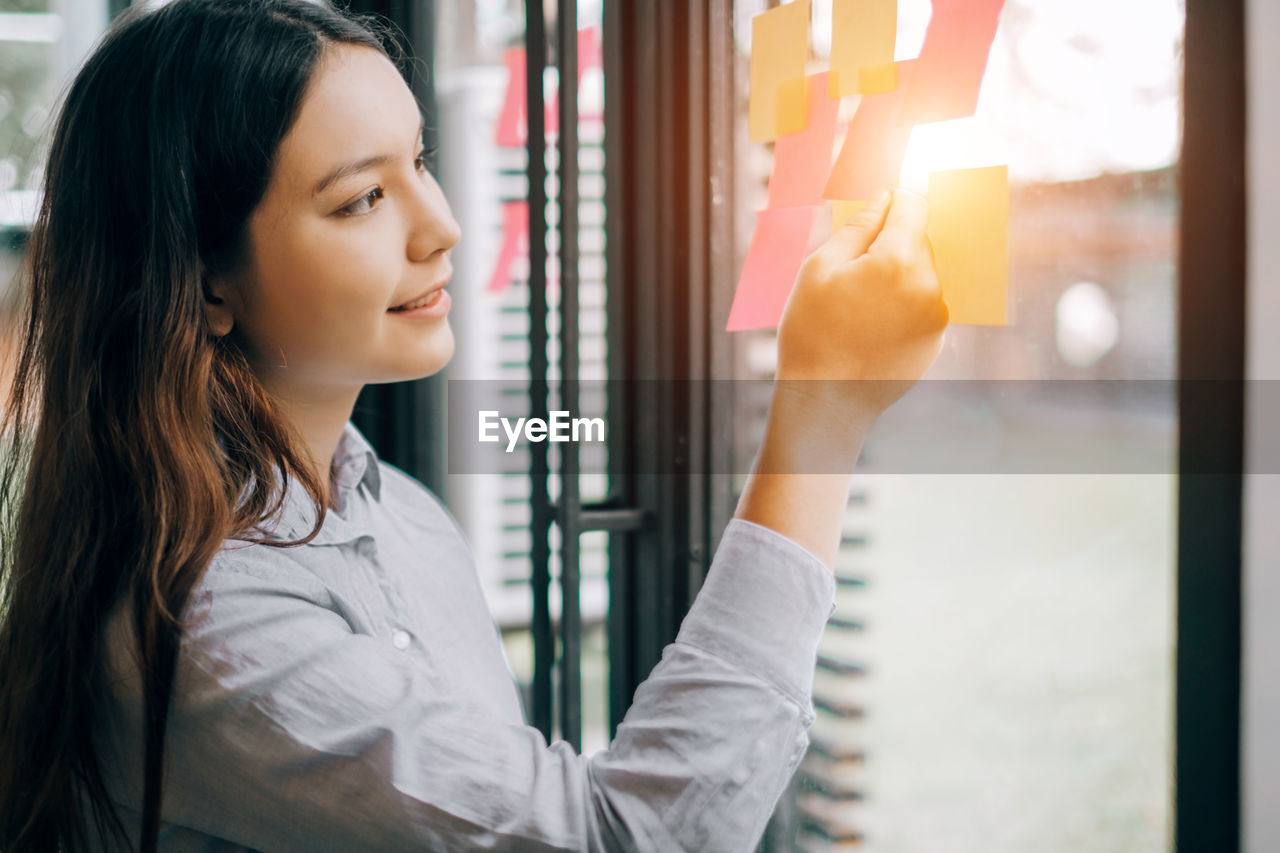 Young businesswoman sticking adhesive note on window at office