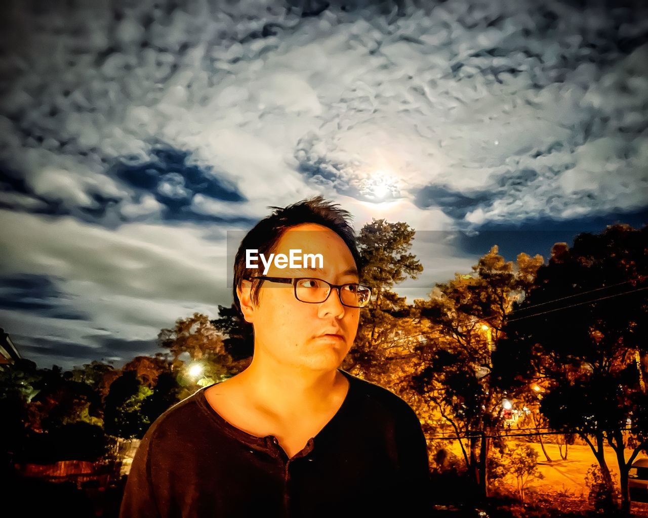 Portrait of young man standing against cloudy sky and rainbow colored lunar halo at night.