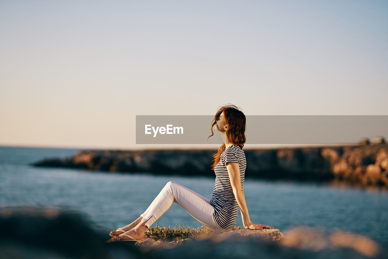 WOMAN SITTING BY SEA AGAINST SKY