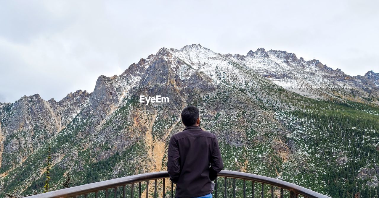 REAR VIEW OF MAN STANDING BY RAILING AGAINST MOUNTAIN