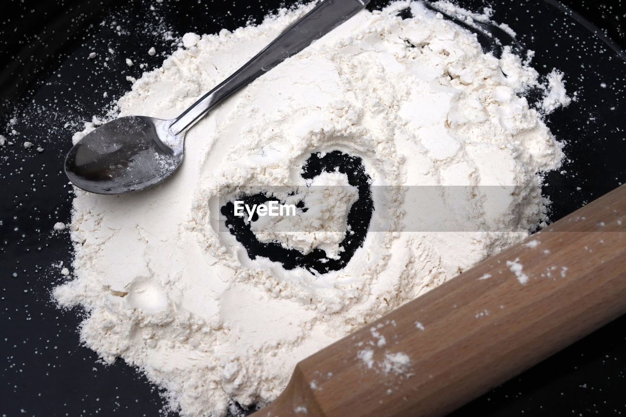 A staging of a heart drawn in flour with a tablespoon and a wooden rolling pin on a black background