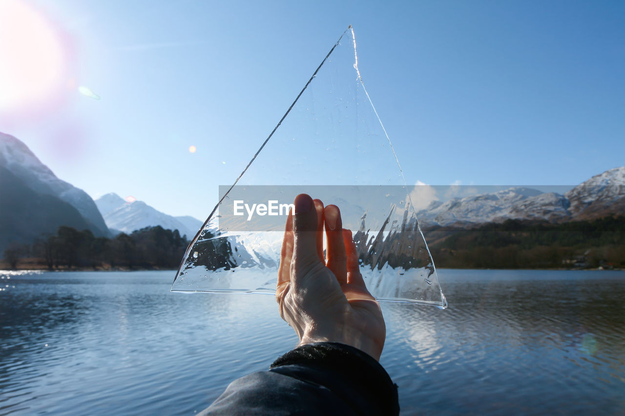 Cropped hand holding ice over river against clear blue sky