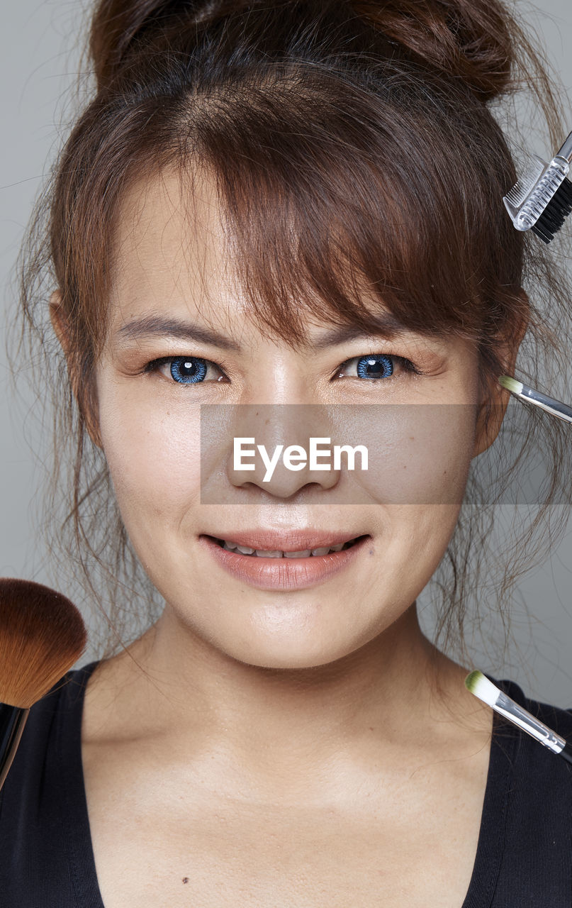 Close-up portrait of smiling beautiful woman with various make-up brushes and mascara applicator against gray background