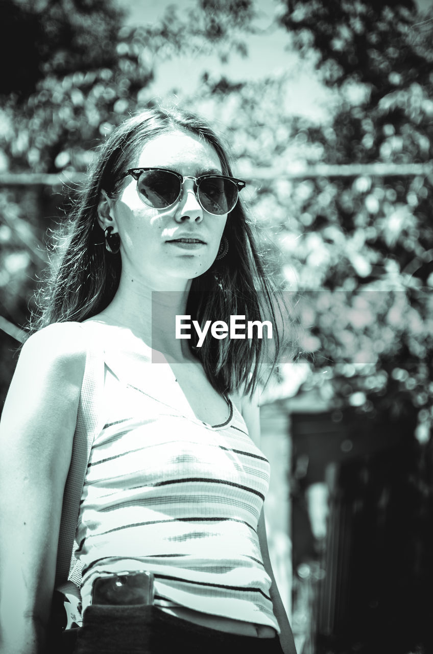 Portrait of young woman wearing sunglasses while standing outdoors during sunny day