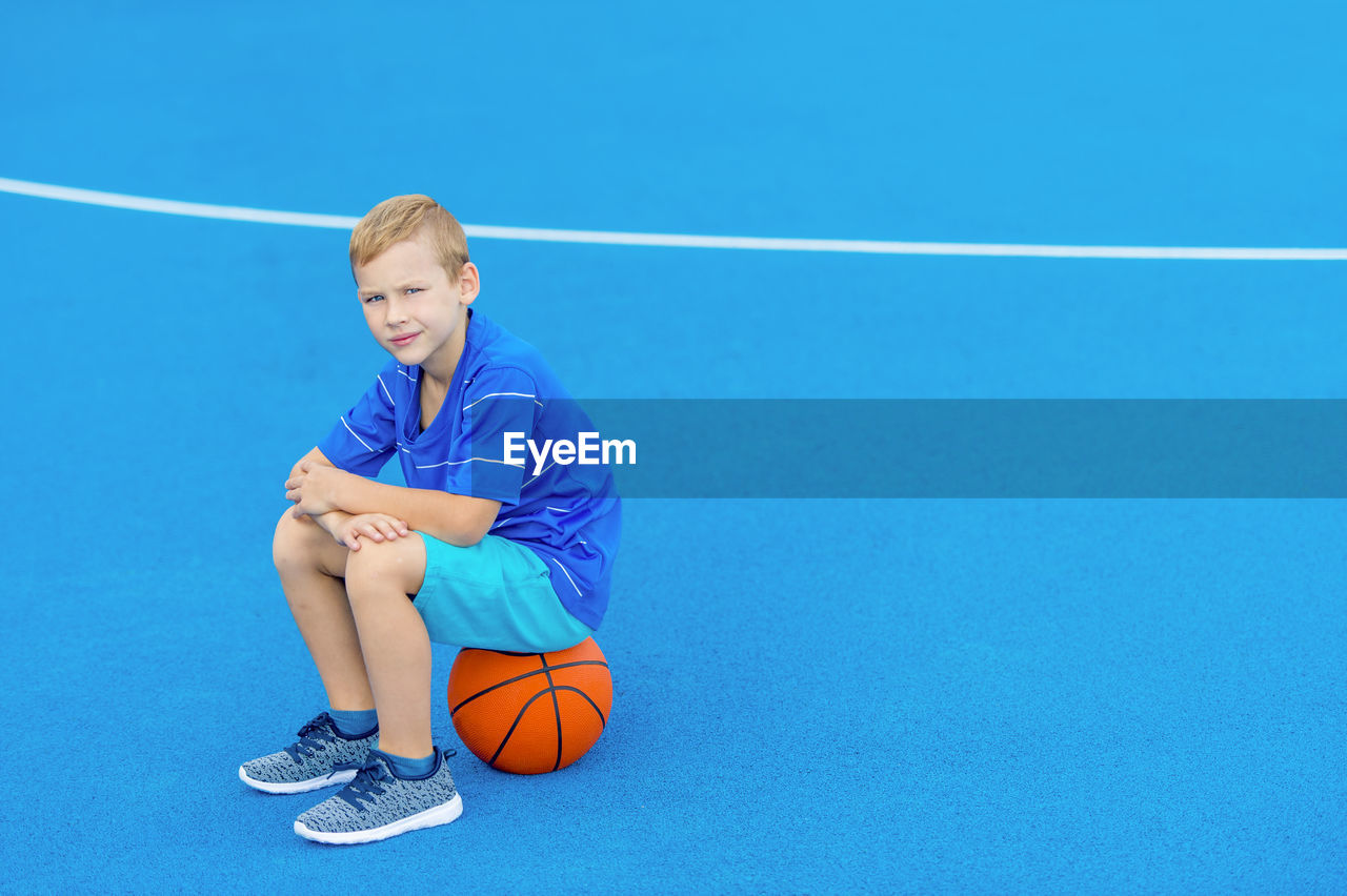 Portrait of boy sitting on basketball at court