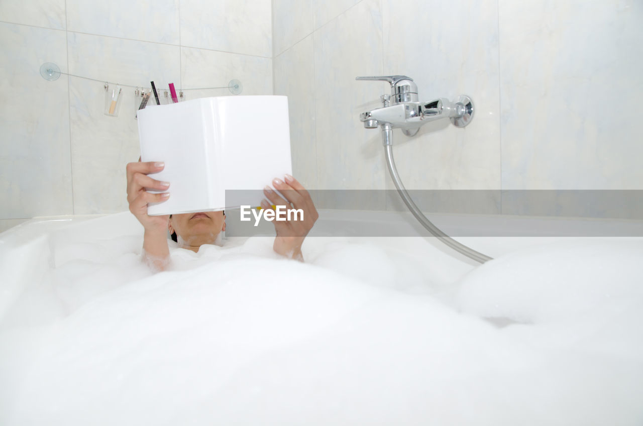 Woman lying in bathtub while reading book