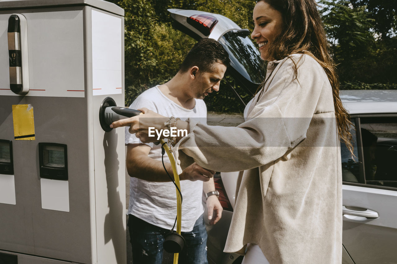 Smiling mother holding electric plug standing by father at charging station