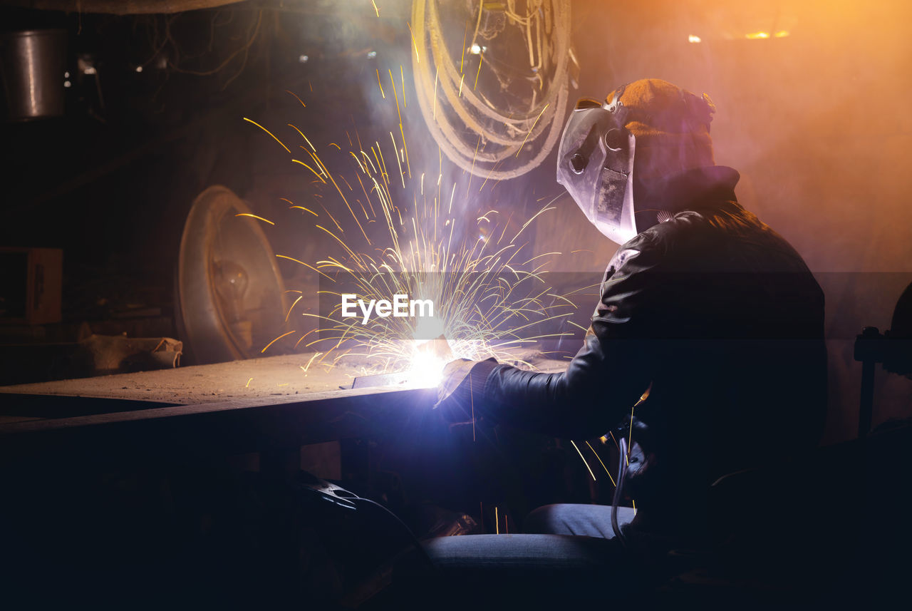 Young mechanic with a welding machine in an old dirty garage at night. man in protection doing