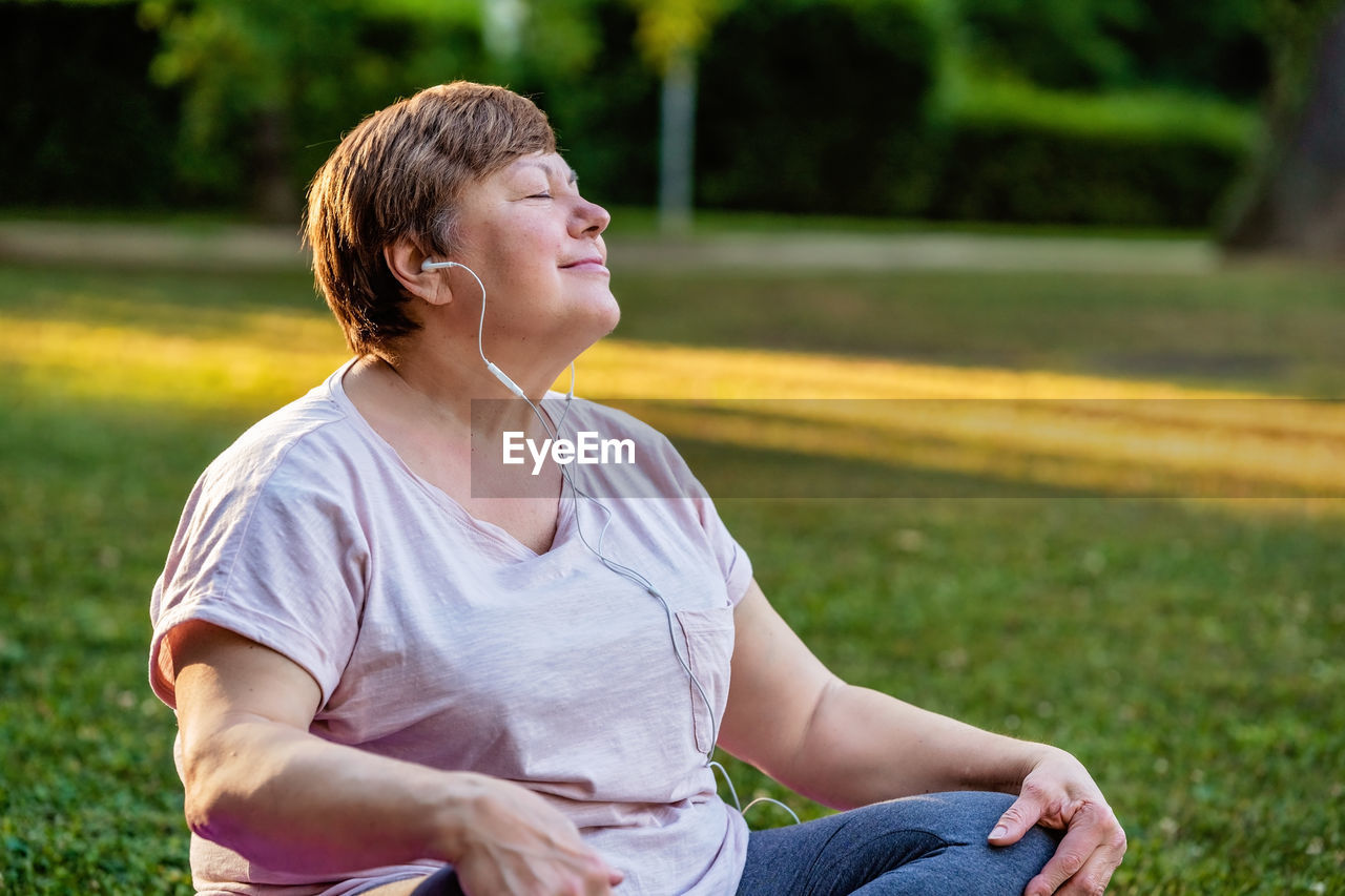 Portrait of relaxed senior plus size woman with earphones sitting with closed eyes on grass in park 