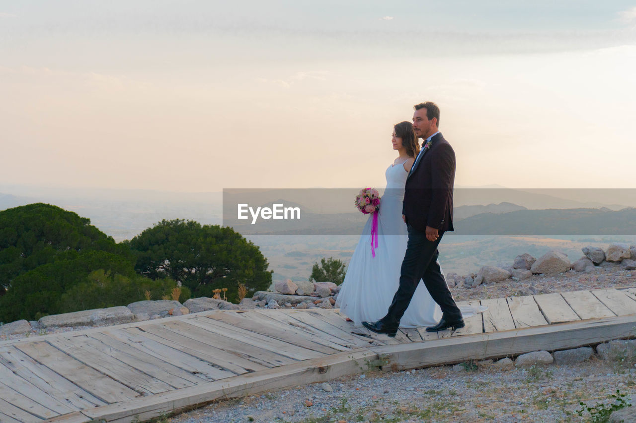 Side view of bride and groom walking on boardwalk against sky