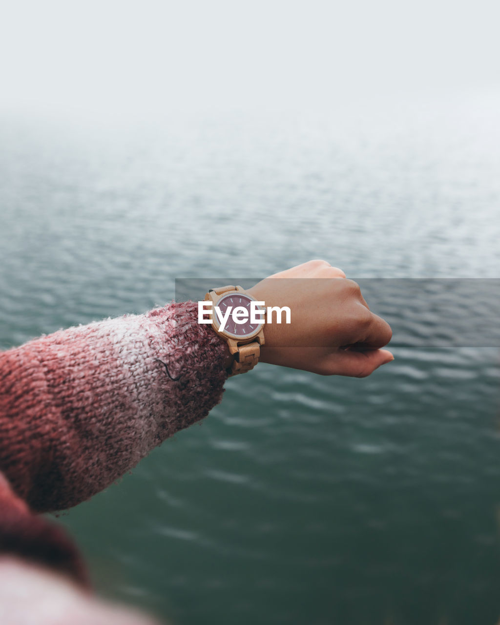 From above crop anonymous female in warm sweater checking time on stylish wristwatch while holding hand over river in cloudy day