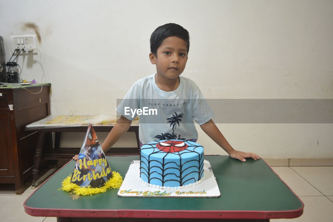 one person, child, childhood, indoors, birthday cake, table, front view, men, art, portrait, person, looking at camera, waist up, sitting, lifestyles, casual clothing, standing, toddler, cake, desk, innocence, clothing, home interior, creativity, domestic life, food, emotion