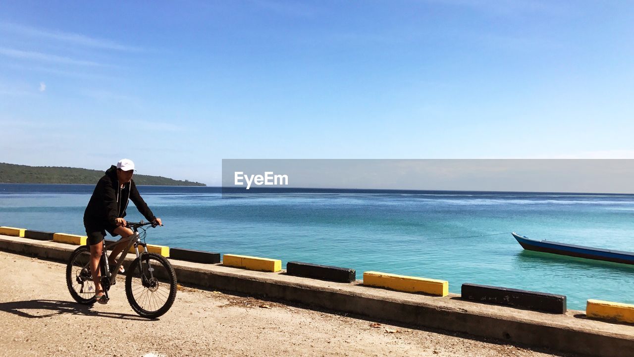 Man riding bicycle on road by sea against sky