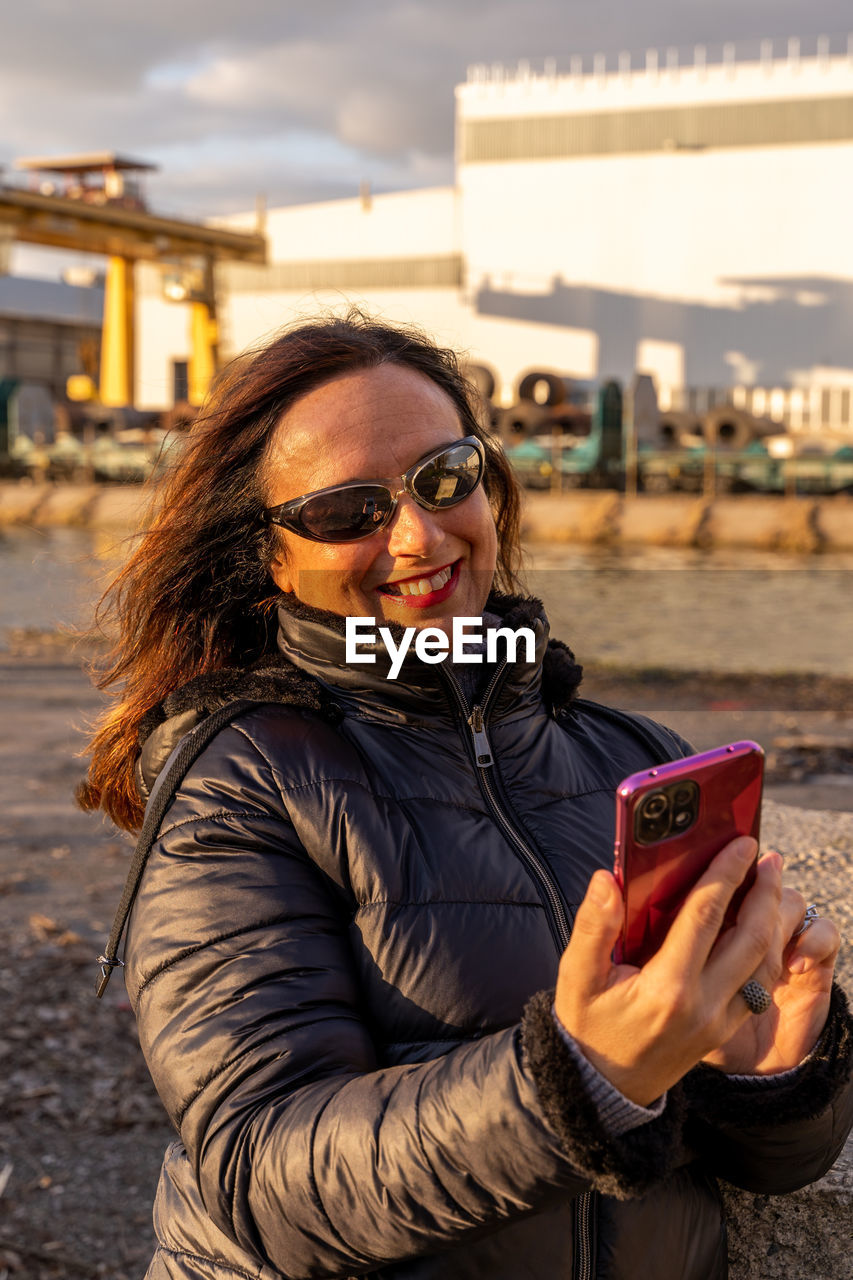 Middle aged woman wearing winter clothes using smartphone in front of a factory