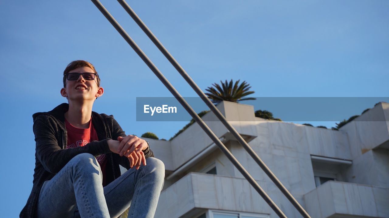 Low angle view of young man wearing sunglasses while sitting on building terrace against clear sky