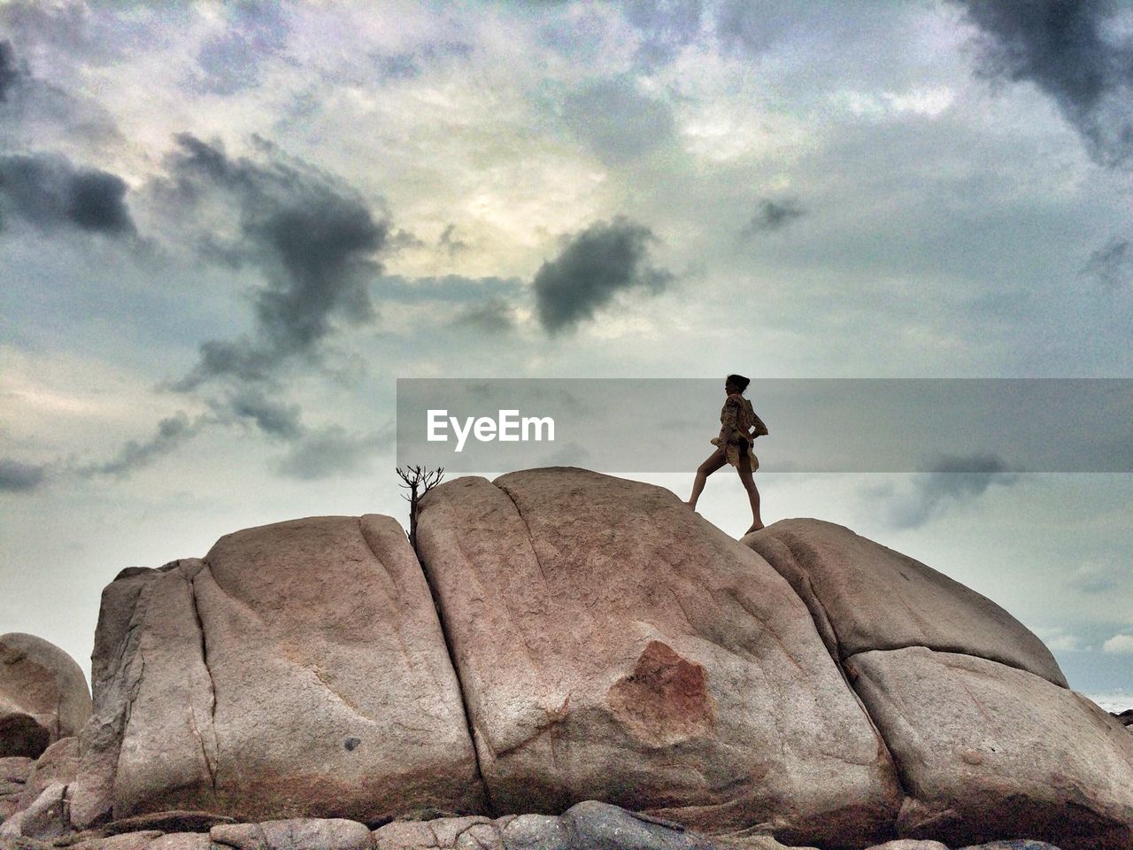 Low angle view of sensuous woman standing on rock against cloudy sky