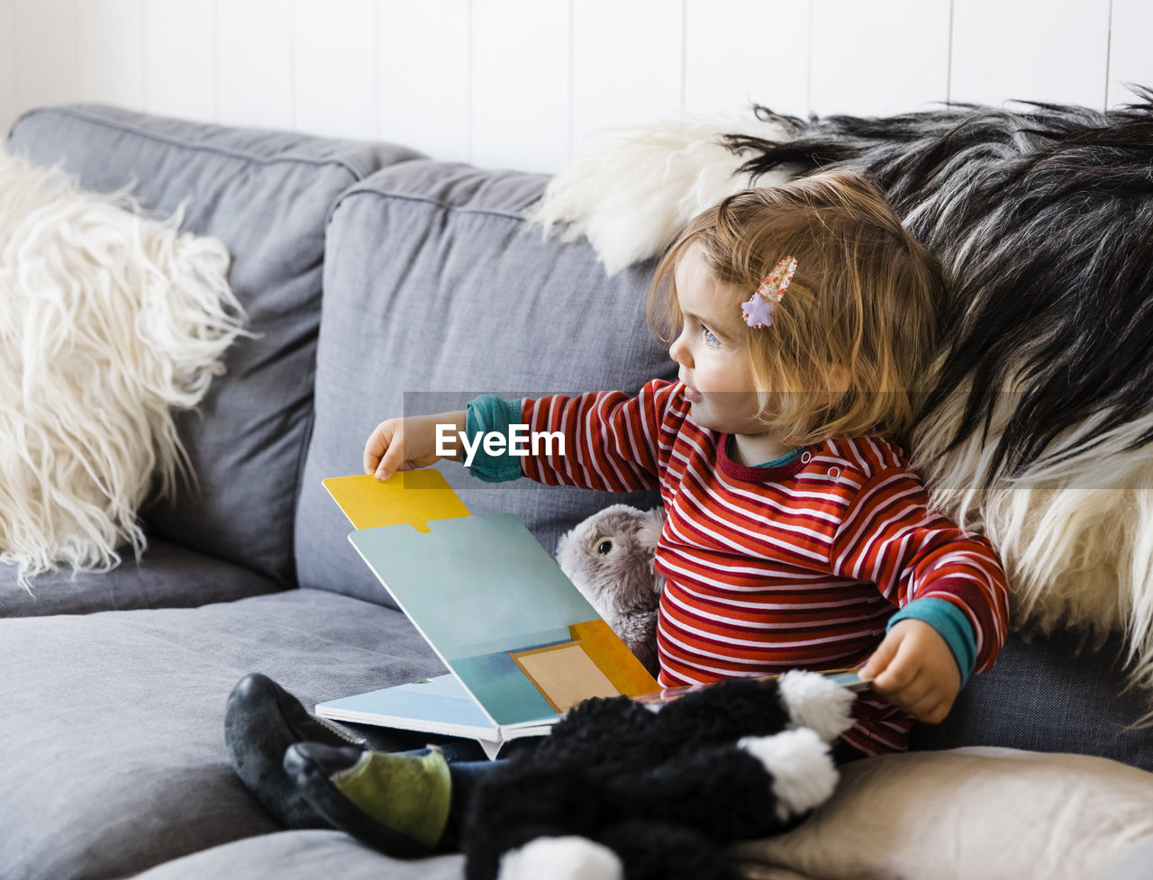 Full length of cute girl with book looking away while sitting on sofa at home