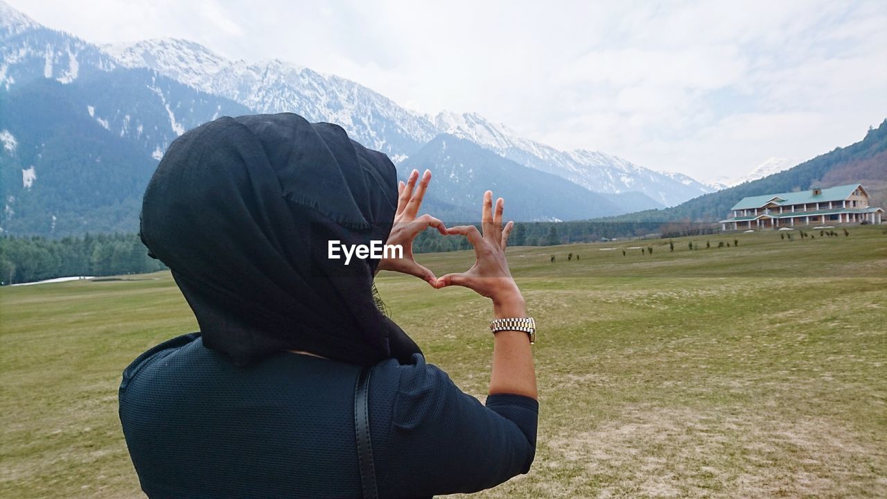 Rear view of woman making heart shape from hands on field against sky