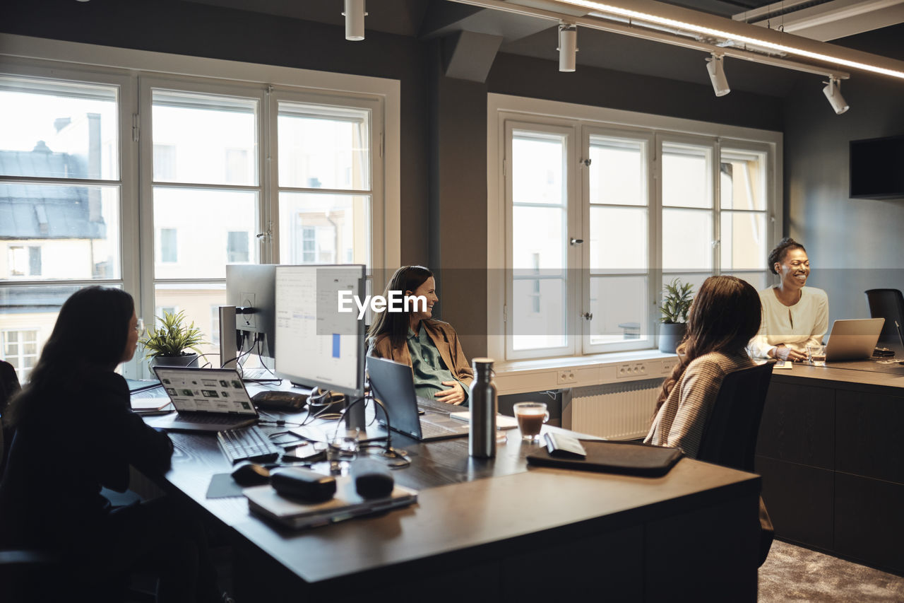 Happy female entrepreneurs communicating with each other while sitting at desk in office