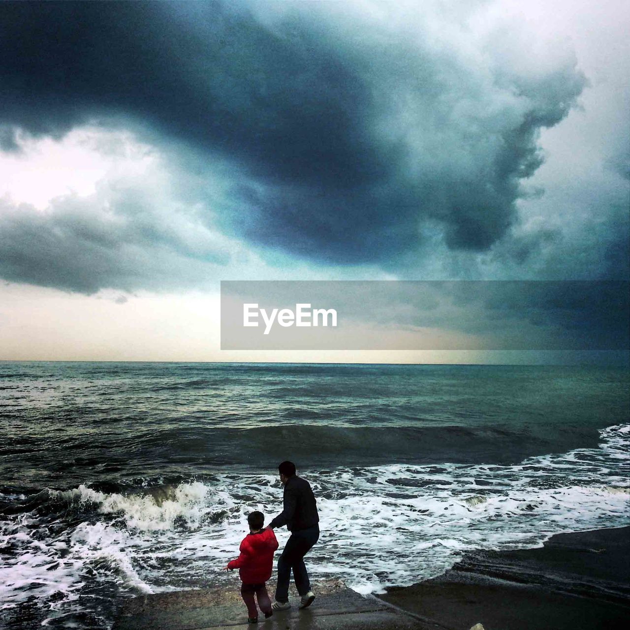 Rear view of father and son at shore against cloudy sky