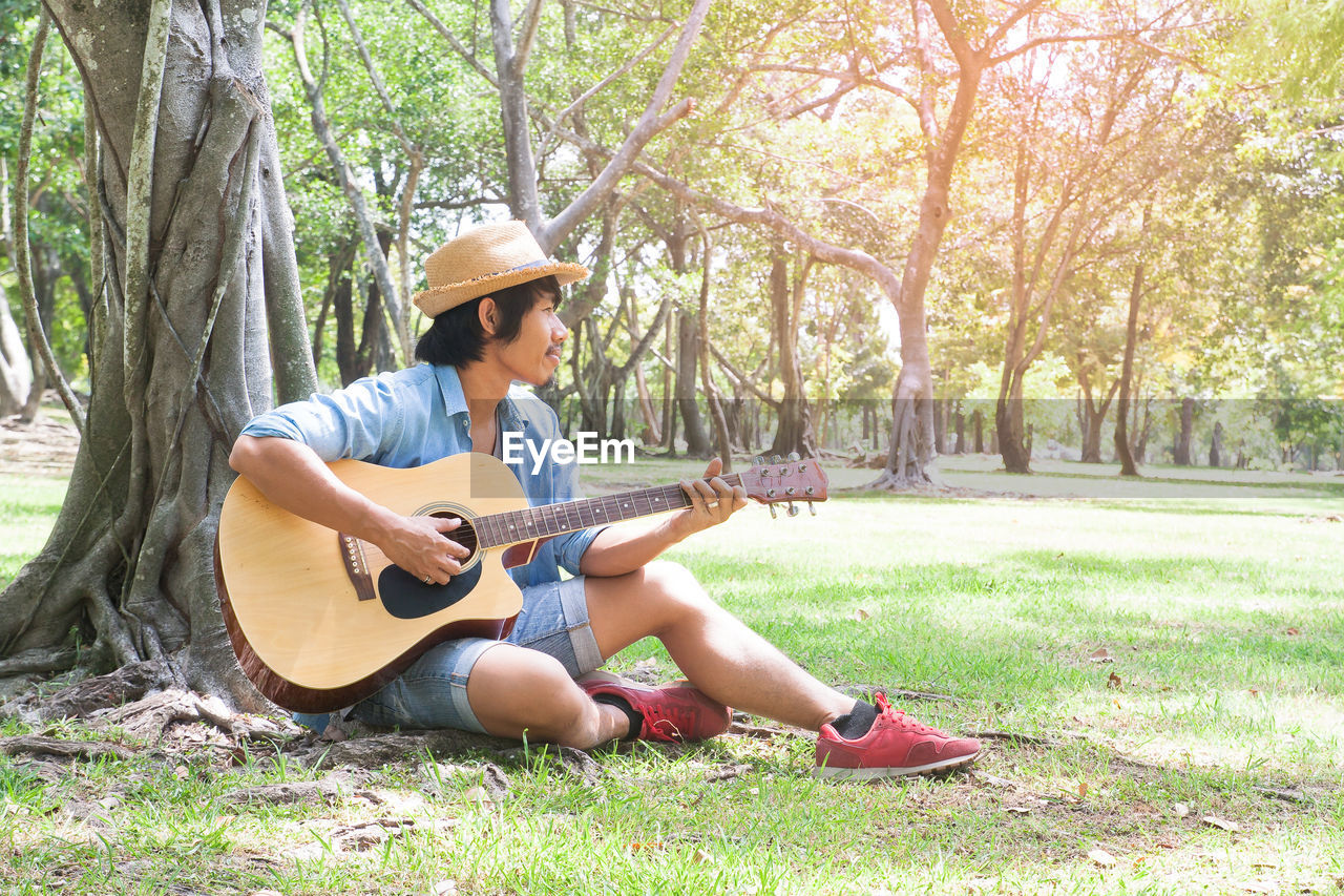 Young man playing guitar while sitting under tree on field at park during sunny day