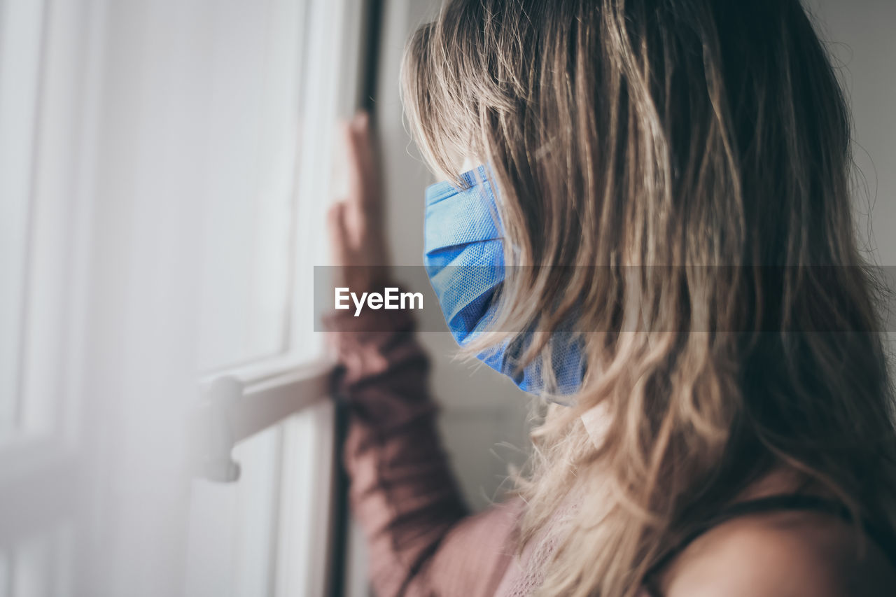 Young woman looking outside window with surgical mask during coronavirus lockdown covid-19 isolated