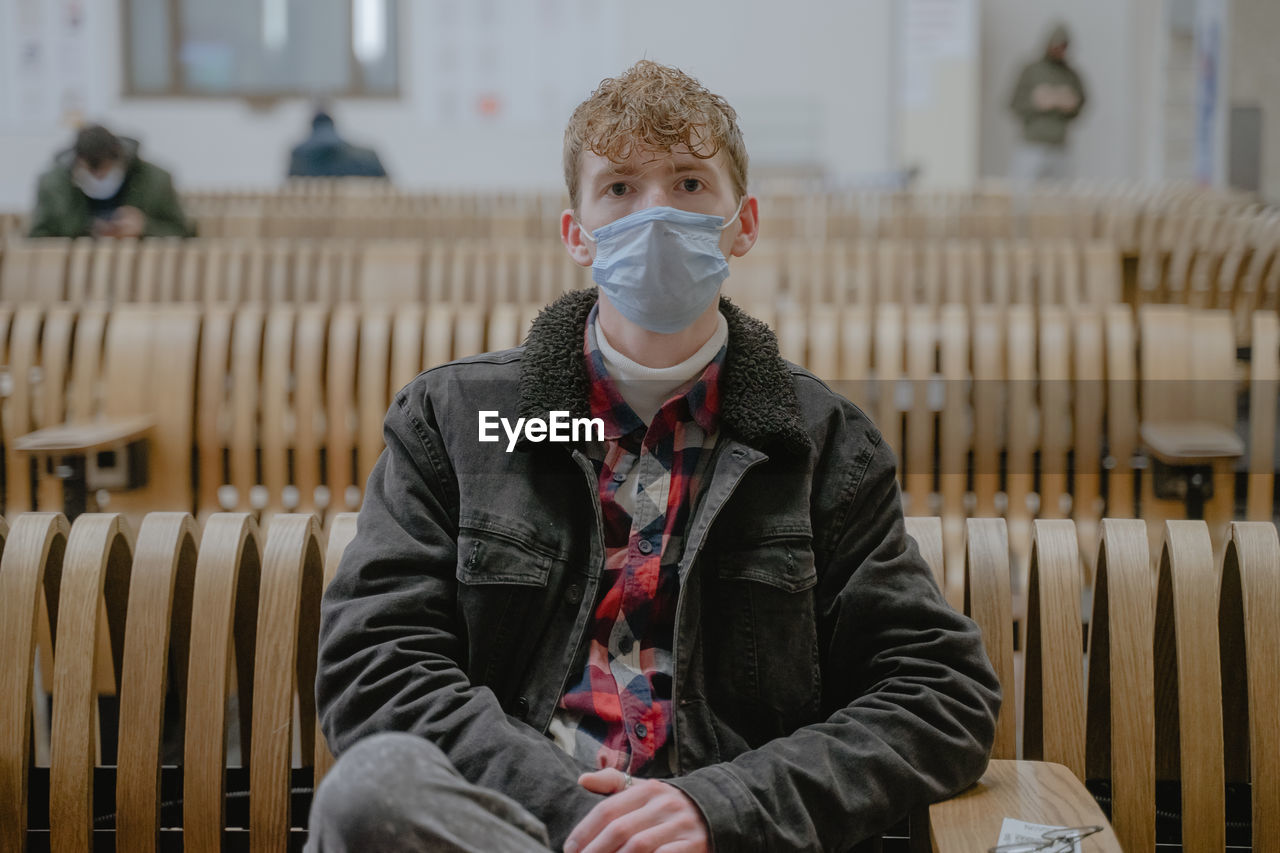 A young man sits at the station waiting for his train, with a mask on his face, curly hair, movement