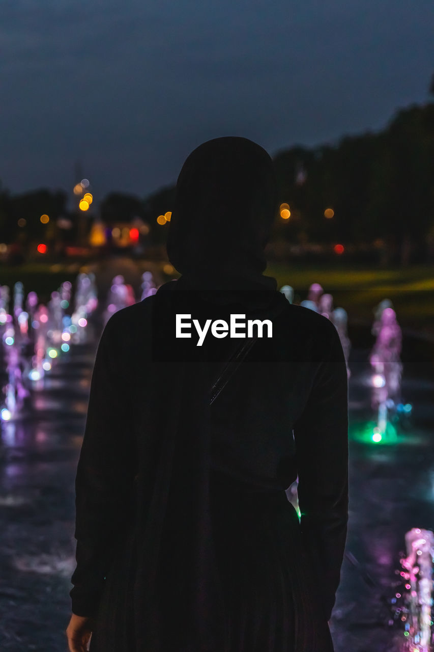 Rear view of muslim woman standing against illuminated city at night