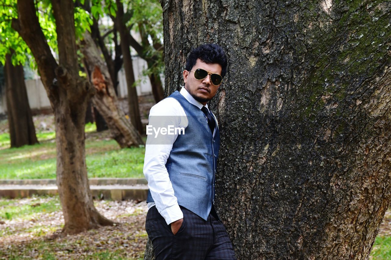 Portrait of young man wearing waistcoat while leaning on tree at park