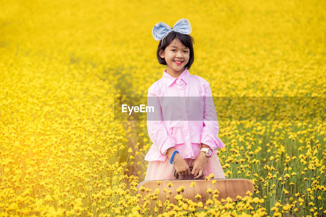Portrait of smiling girl with case standing amidst yellow flowers on field