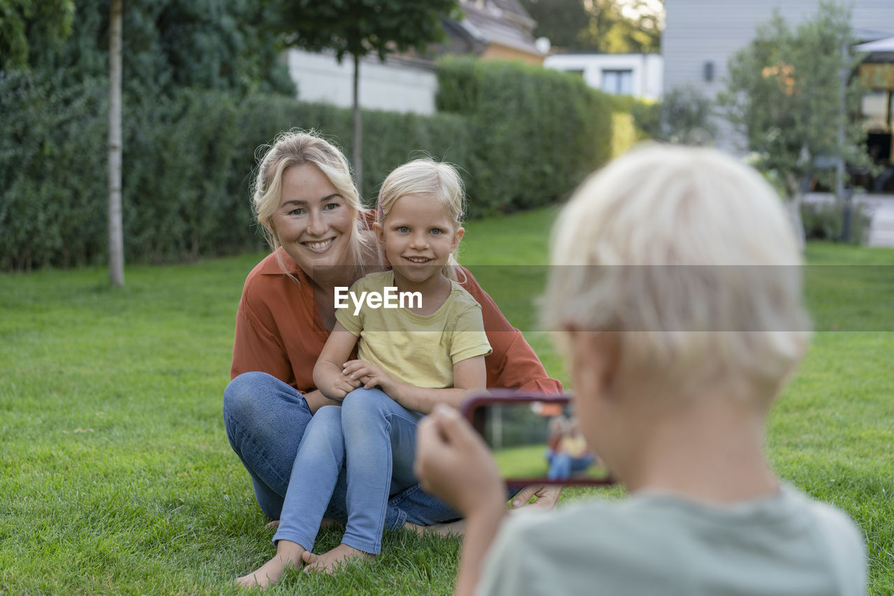 Boy photographing smiling mother and sister through smart phone in garden