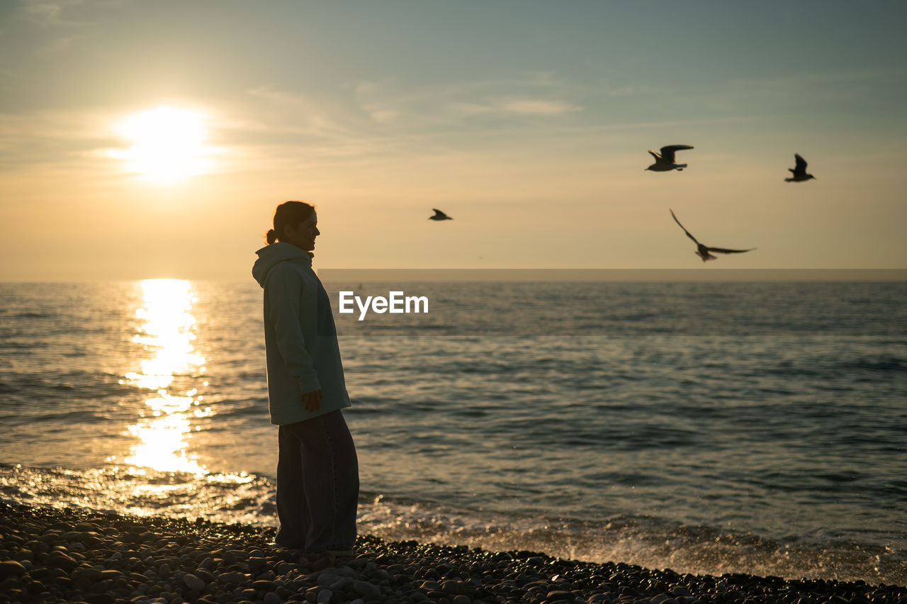 rear view of silhouette woman standing at beach against sky during sunset