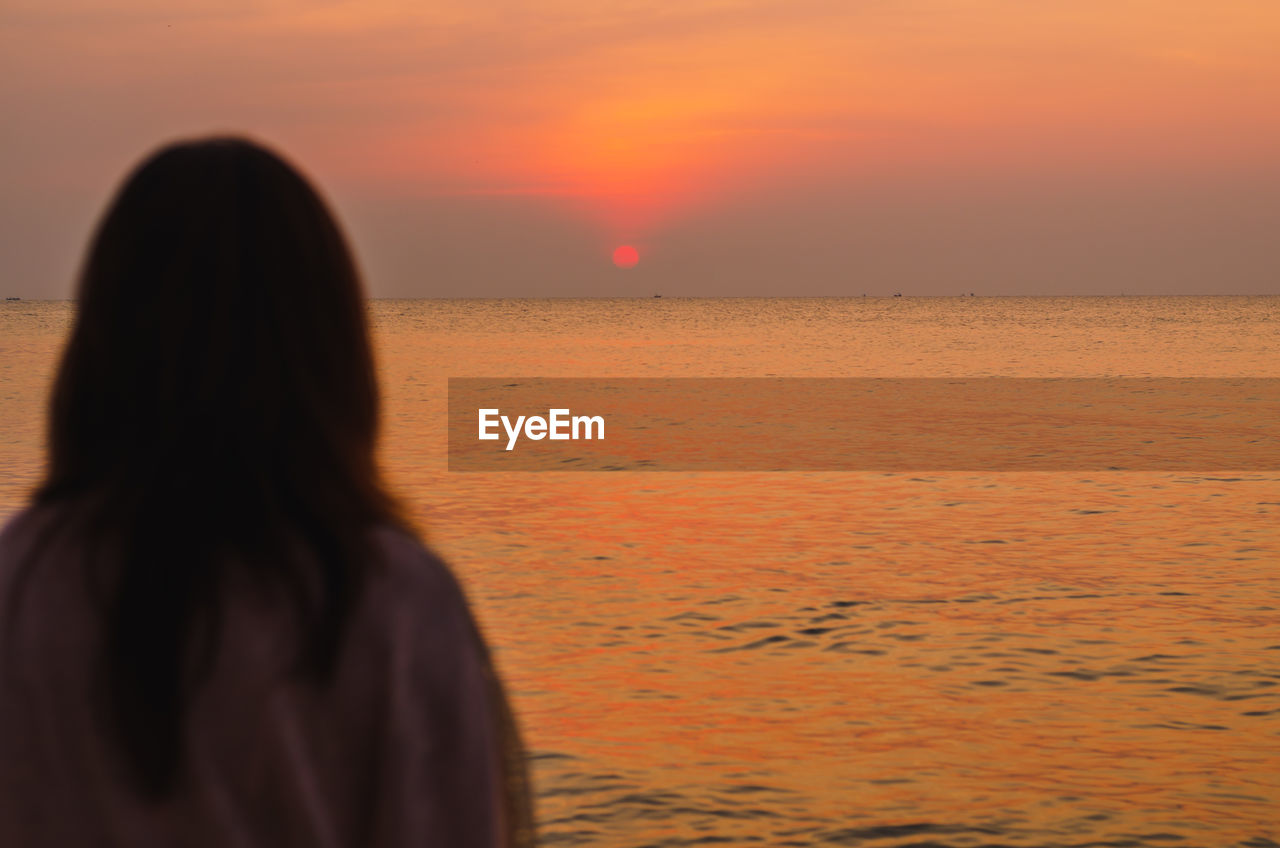 REAR VIEW OF WOMAN STANDING ON BEACH AGAINST SKY DURING SUNSET