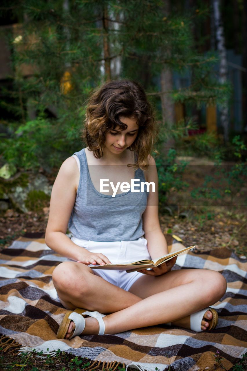 Beautiful teenager girl reading a book lying on blanket on green grass at the woods.