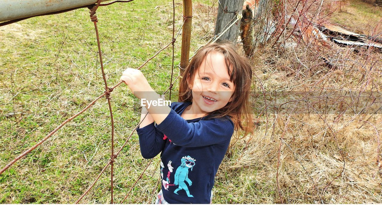 Portrait of smiling cute girl holding fence on grassy land