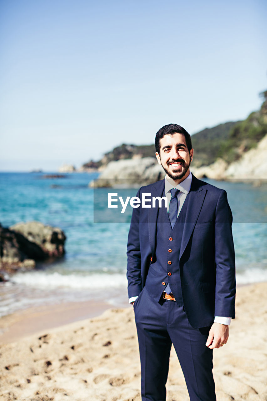 Portrait of smiling bridegroom standing at beach against clear sky during sunny day