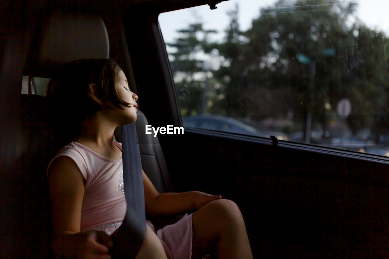 A beautiful little girl wearing seat belt sleeps in a car at twilight