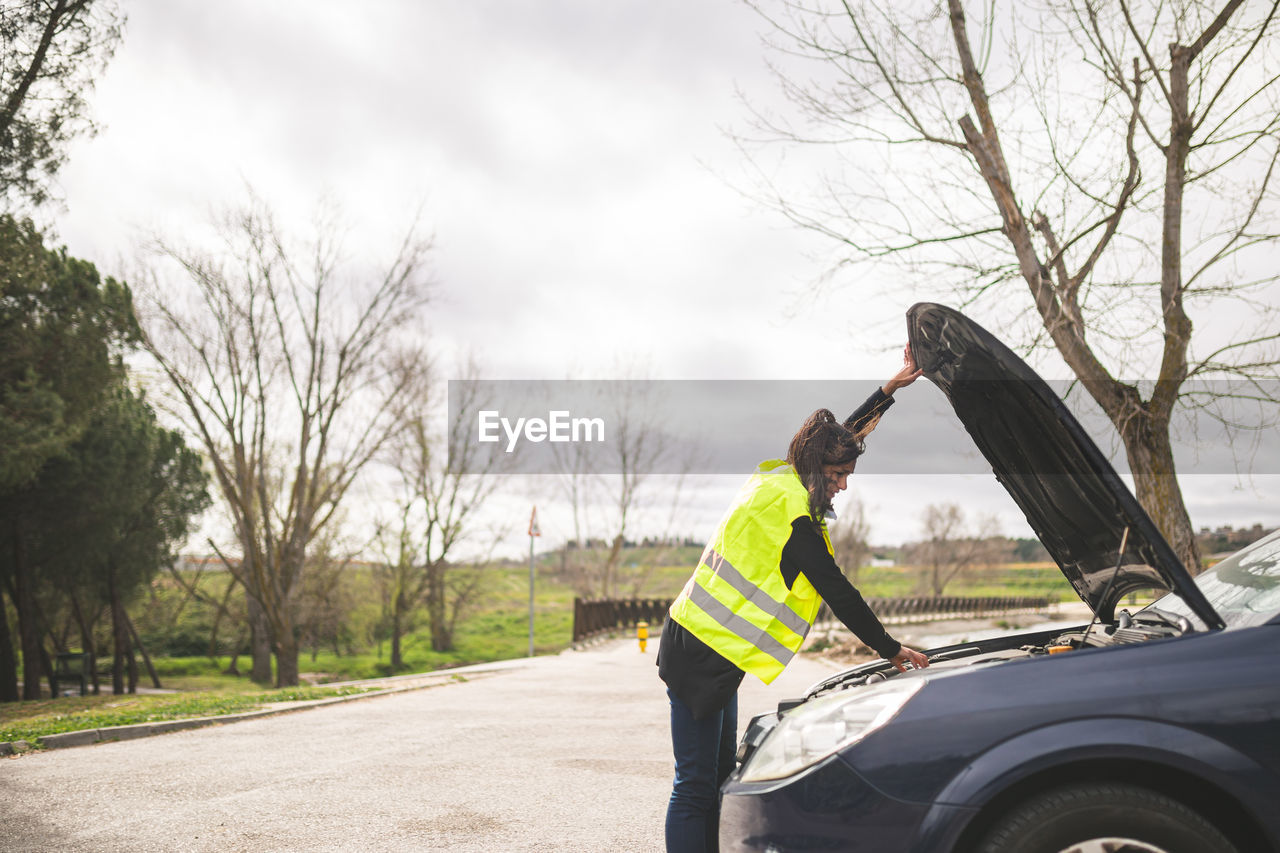 Young caucasian woman looking at car engine, car broken down in the middle of the road. 