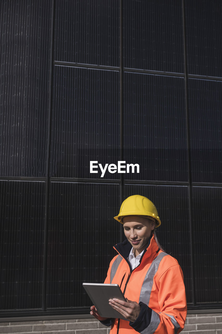 Young female engineer wearing hardhat using tablet pc in front of solar panels