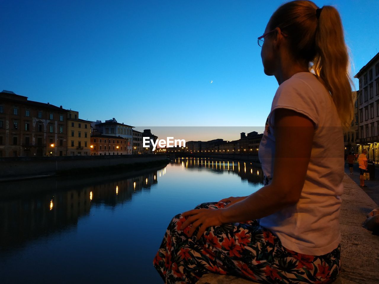 Side view of woman sitting by canal in city against sky