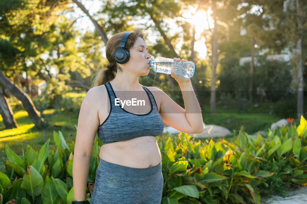 side view of woman drinking water while standing against trees