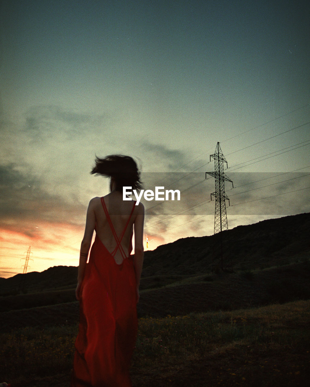 REAR VIEW OF WOMAN STANDING ON FIELD AGAINST SKY DURING SUNSET